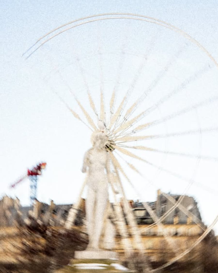 Une statue blanche devant une grande roue floue, sur fond de ciel pâle.