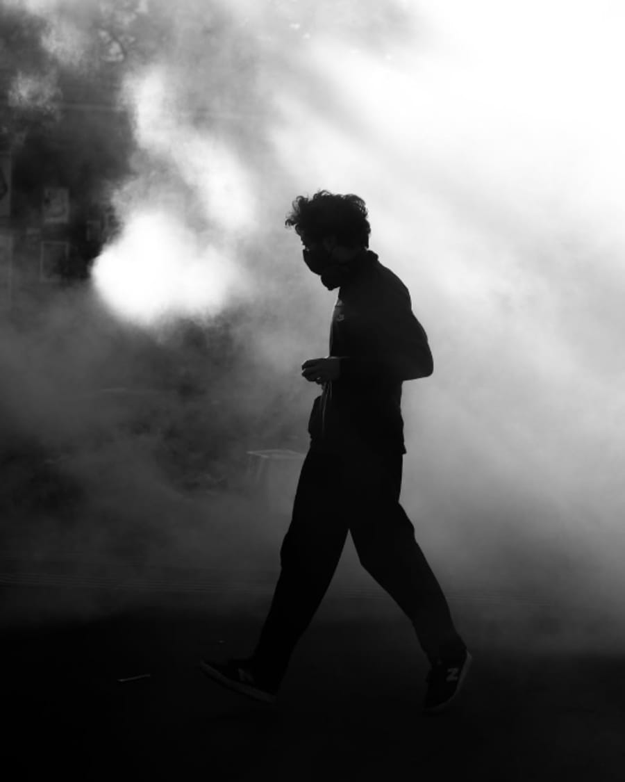 La silhouette d'un passant masqué qui marche dans une brume traversée de lumière.